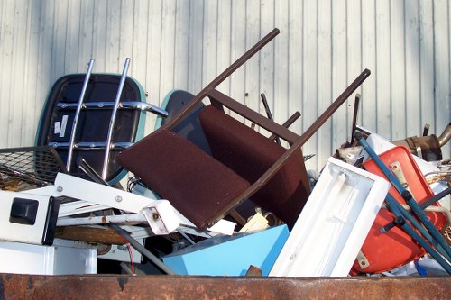 A large pile of discarded household items and rubbish stacked outdoors, including a burgundy upholstered armchair with wooden arms, a white domestic fridge, a blue plastic storage container, a red plastic child's chair, and various metal and plastic objects. The debris is positioned on a concrete or tarmac surface against a background of light grey, vertically ribbed metal siding. The items appear to be part of a house clearance, with some objects tilted or leaning, and the scene suggests an ongoing rubbish removal process typical of house clearance services around Welwyn area. The lighting is diffuse, with no sharp shadows, indicating a cloudy day, and the overall composition emphasizes the variety of household waste accumulated in preparation for disposal or recycling, as handled by companies like House Clearance Welwyn.