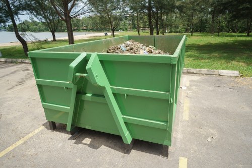 A large, green metal skip situated in a paved outdoor parking area near a grassy park with trees and a pond visible in the background. The skip is filled with various types of refuse, including cardboard, plastic, and miscellaneous household waste. The surface of the skip has a textured, ribbed appearance, and it is positioned with its open top revealing the contents inside. Surrounding the skip are faint yellow painted lines indicating designated parking spaces, and the overall environment appears clean and orderly. The scene is well-lit with natural daylight, emphasizing the contrast between the greenery of the park and the utilitarian appearance of the skip, which is managed by a rubbish removal service focused on clearing waste amidst a quiet suburban setting.