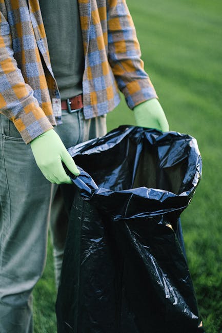 A pile of mixed household rubbish is scattered on a gravel surface in an outdoor area, with black plastic trash bags, discarded cardboard and paper, and a broken, dirty beige upholstered chair with wo