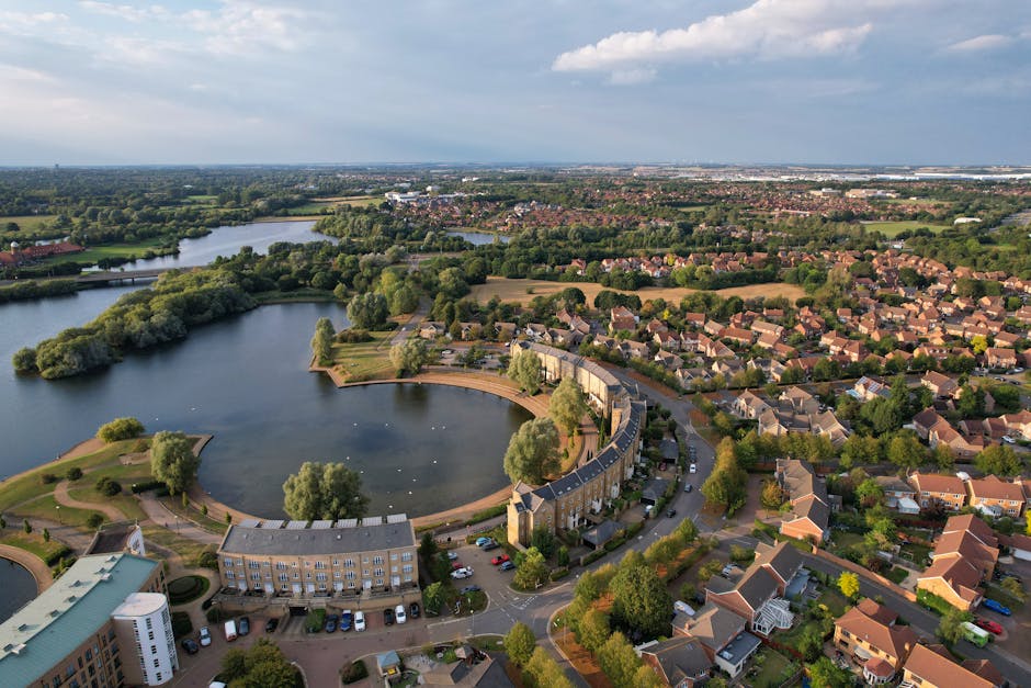 An aerial view of a residential area featuring a large, curved lake with a concrete promenade along its edge, surrounded by numerous trees and green spaces. The lake has a smooth surface with a few sm